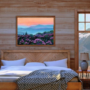 May include: A rectangular landscape painting in a wooden frame depicting a field of vibrant purple rhododendron flowers in the foreground with rolling blue mountain ridges under a soft peach and orange sunset sky, signed by the artist in the corner.