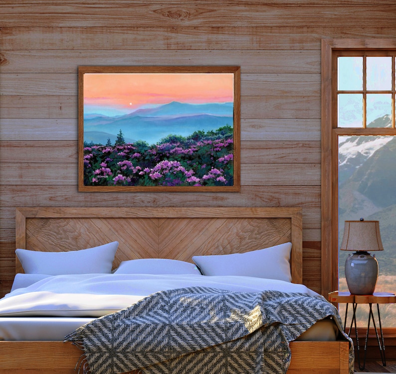 May include: A rectangular landscape painting in a wooden frame depicting a field of vibrant purple rhododendron flowers in the foreground with rolling blue mountain ridges under a soft peach and orange sunset sky, signed by the artist in the corner.