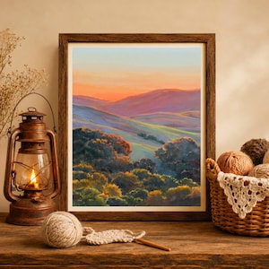 May include: A framed landscape painting depicting rolling hills at sunset with orange and purple skies, positioned next to a rustic metal lantern with a glowing flame and a woven basket filled with balls of yarn and a crochet hook on a wooden surface.