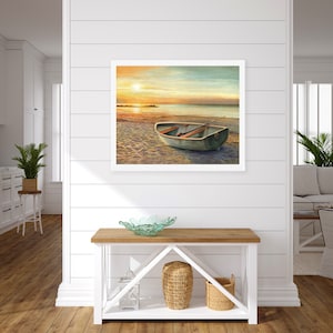 Op de afbeelding: Een ingelijst landschapsschilderij met een kleine, lichtgekleurde roeiboot op een zandstrand bij zonsondergang. De sc&egrave;ne toont een kalme oceaanhorizon onder een warme, gouden lucht, met de boot centraal in de compositie geplaatst.