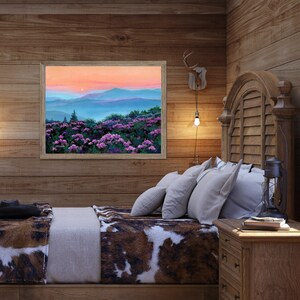 May include: A framed landscape painting depicting a sunset over blue mountain ranges with a foreground of vibrant purple flowers. The artwork features a soft orange and pink sky, layered blue hills, and dense green foliage with purple blossoms.