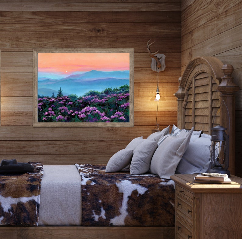 May include: A framed landscape painting depicting a sunset over blue mountain ranges with a foreground of vibrant purple flowers. The artwork features a soft orange and pink sky, layered blue hills, and dense green foliage with purple blossoms.