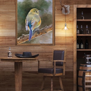 May include: A framed wall art print featuring a small bird with yellow, green, and blue plumage perched on a textured tree branch. The artwork is displayed on a wooden wall above a round wooden table with a black hat and a small glass bottle.