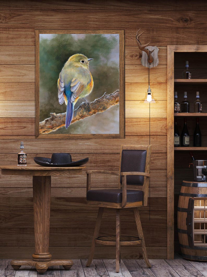 May include: A framed wall art print featuring a small bird with yellow, green, and blue plumage perched on a textured tree branch. The artwork is displayed on a wooden wall above a round wooden table with a black hat and a small glass bottle.