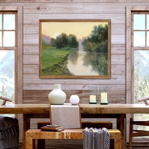 Pode incluir: Uma pintura de paisagem emoldurada retratando um rio que flui atrav&eacute;s de um prado verde com &aacute;rvores sob um c&eacute;u suave de p&ocirc;r do sol. A obra de arte &eacute; exibida em uma parede de pain&eacute;is de madeira clara acima de uma mesa de jantar de madeira.
