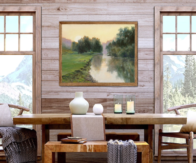 Pode incluir: Uma pintura de paisagem emoldurada retratando um rio que flui atrav&eacute;s de um prado verde com &aacute;rvores sob um c&eacute;u suave de p&ocirc;r do sol. A obra de arte &eacute; exibida em uma parede de pain&eacute;is de madeira clara acima de uma mesa de jantar de madeira.