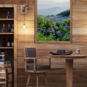 May include: A framed landscape painting depicting rolling blue mountains behind a foreground of vibrant green foliage and pink flowers. The artwork is displayed on a wood-paneled wall above a wooden swivel chair and a round pedestal table.