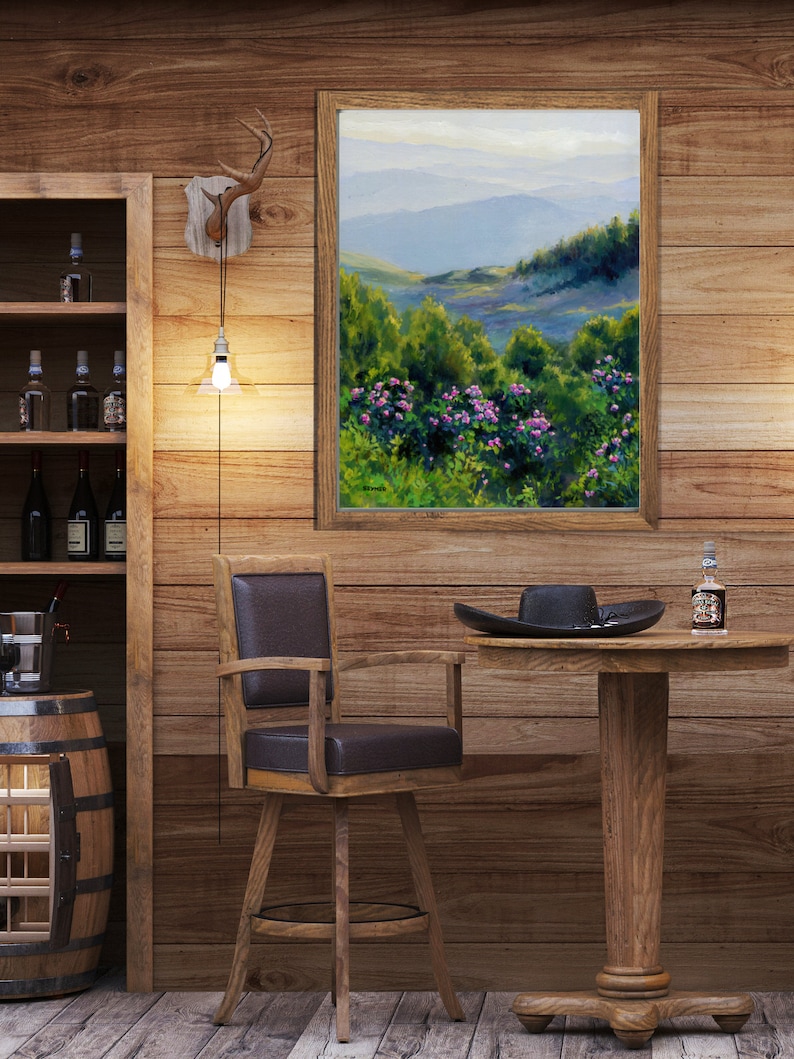 May include: A framed landscape painting depicting rolling blue mountains behind a foreground of vibrant green foliage and pink flowers. The artwork is displayed on a wood-paneled wall above a wooden swivel chair and a round pedestal table.