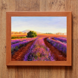 May include: A painting of a lavender field with a winding dirt road leading to a small grove of trees in the distance. The sky is a soft blue with hints of pink and orange. The painting is framed in a light brown wood frame.