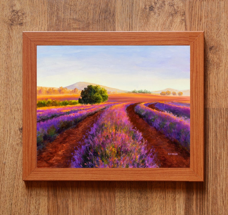 May include: A painting of a lavender field with a winding dirt road leading to a small grove of trees in the distance. The sky is a soft blue with hints of pink and orange. The painting is framed in a light brown wood frame.