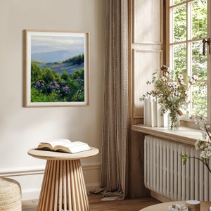 May include: A square framed landscape painting hanging on a wall, depicting a mountain range in the distance with rolling green hills and vibrant purple flowers in the foreground, rendered in an oil-on-canvas style with visible brushstrokes.