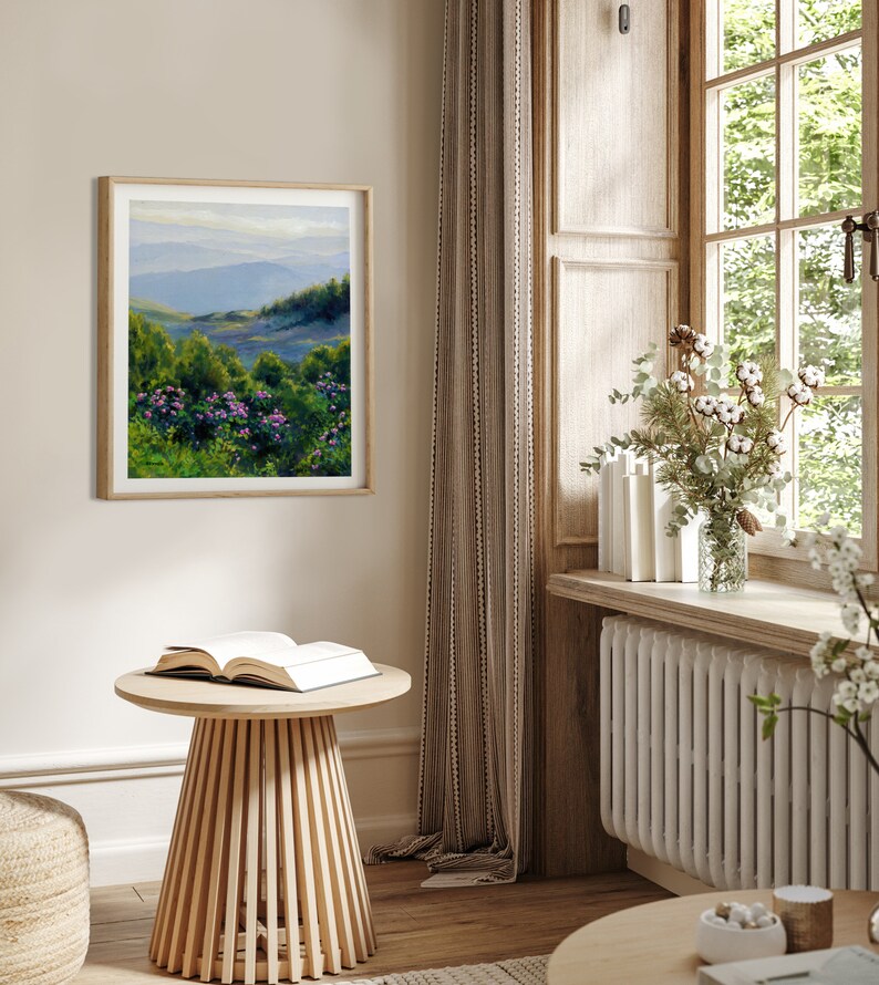 May include: A square framed landscape painting hanging on a wall, depicting a mountain range in the distance with rolling green hills and vibrant purple flowers in the foreground, rendered in an oil-on-canvas style with visible brushstrokes.