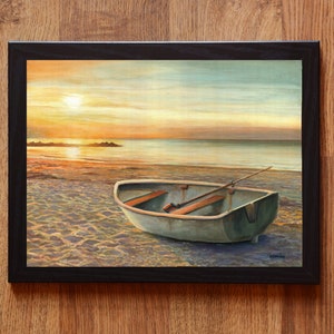 Op de afbeelding: Een ingelijst schilderij met een kleine, lichtgekleurde roeiboot op een zandstrand bij zonsondergang. De sc&egrave;ne toont een kalme oceaan, een felle zon laag aan de horizon en een enkele houten roeispaan in de boot. De lijst is zwart.