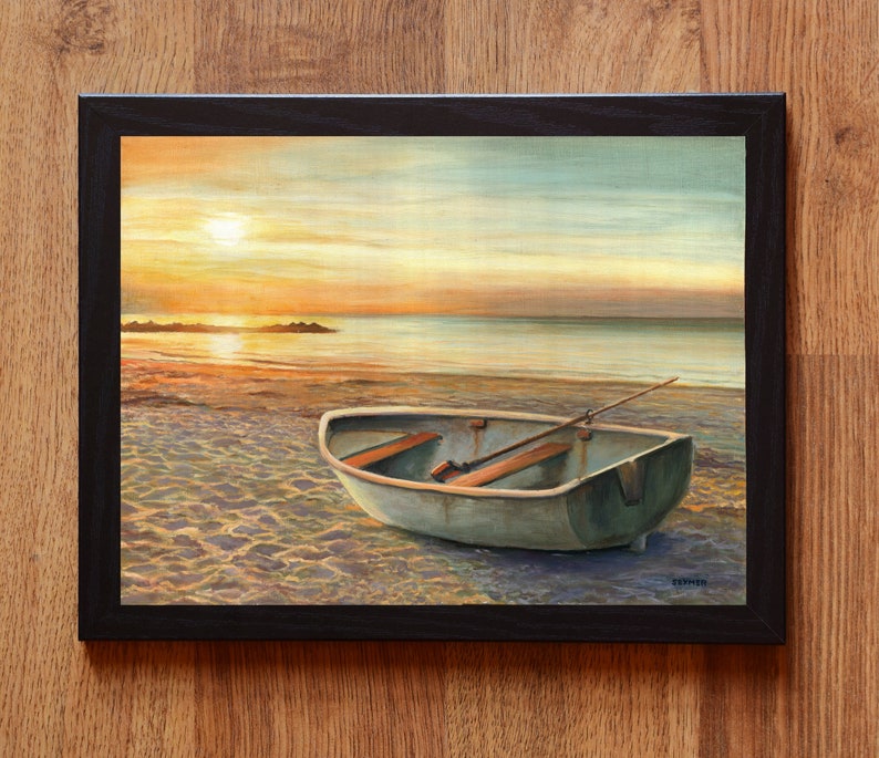 Op de afbeelding: Een ingelijst schilderij met een kleine, lichtgekleurde roeiboot op een zandstrand bij zonsondergang. De sc&egrave;ne toont een kalme oceaan, een felle zon laag aan de horizon en een enkele houten roeispaan in de boot. De lijst is zwart.