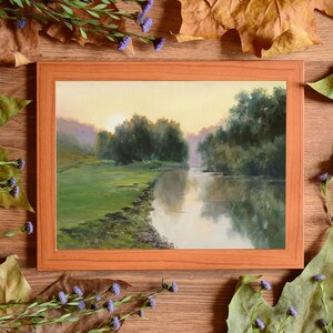 Pode incluir: Uma pintura de paisagem retangular numa moldura de madeira castanho-claro que retrata um rio a fluir atrav&eacute;s de um prado verde ao nascer do sol. A cena apresenta &aacute;rvores verde-escuras ao longo da margem e reflexos na superf&iacute;cie da &aacute;gua.