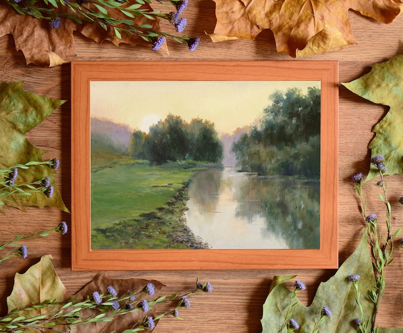 Pode incluir: Uma pintura de paisagem retangular numa moldura de madeira castanho-claro que retrata um rio a fluir atrav&eacute;s de um prado verde ao nascer do sol. A cena apresenta &aacute;rvores verde-escuras ao longo da margem e reflexos na superf&iacute;cie da &aacute;gua.