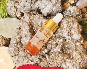 May include: A small glass bottle with a gold cap and a dropper, filled with a clear liquid. The bottle is labeled "manifest". The bottle is sitting on a rock with a red rose in the foreground.