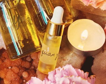 May include: Three glass bottles with gold lids and labels. The bottles contain a clear liquid. The labels have the text "Guan Yin" on them. The bottles are sitting on a surface with a pink flower and a lit candle.