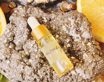 May include: A small, clear glass bottle with a gold cap and dropper. The bottle is filled with a yellow liquid and has a label that says "money". The bottle is sitting on a piece of pyrite, a type of mineral.