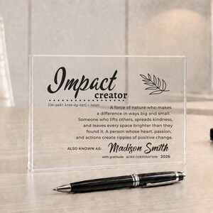 Könnte beinhalten: Eine rechteckige Auszeichnungstafel aus klarem Acryl mit schwarzem Aufdruck, der Impact creator, eine Definition des Begriffs sowie den Namen Madison Smith und das Jahr 2026 zeigt. Ein schwarzer Kugelschreiber liegt horizontal vor der Tafel.