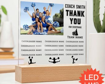 May include: A personalized acrylic plaque with a photo of a cheerleading team in blue and white uniforms. The plaque reads "COACH SMITH THANK YOU FOR EVERYTHING" and includes space for the team name and cheerleader names. It sits on a wooden base with an LED lamp.