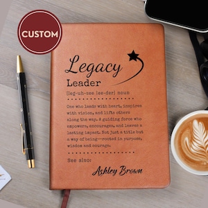 May include: A tan leather-bound journal featuring the black printed text Legacy Leader, a definition of the term, and the name Ashley Brown. A black pen with gold accents rests to the left of the rectangular notebook on a wooden surface.