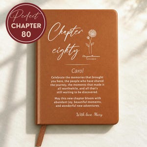 May include: A rectangular brown notebook with a leather-like cover featuring white text that reads Chapter eighty, Carol, and a message about celebrating memories. It includes a line drawing of a chrysanthemum flower and the text With love, Mary.