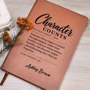 May include: A tan leather-bound journal featuring black printed text that reads Character COUNTS, followed by a definition and the name Ashley Brown. A small sprig of dried orange flowers is positioned on the left side of the journal cover.