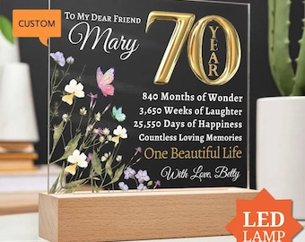 May include: A rectangular clear acrylic plaque with a black background, featuring gold text and a floral design with butterflies. The text reads: To My Dear Friend Mary 70, 840 Months of Wonder, 3,650 Weeks of Laughter, 25,550 Days of Happiness, Countless Loving Memories, One Beautiful Life, With Love, Betty. The plaque is set in a light-colored wooden base.