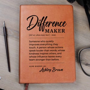 May include: A tan leather-bound notebook featuring the black printed text "Difference MAKER" followed by a definition and the name "Ashley Brown". A black fountain pen is positioned to the left of the rectangular notebook on a light wood surface.
