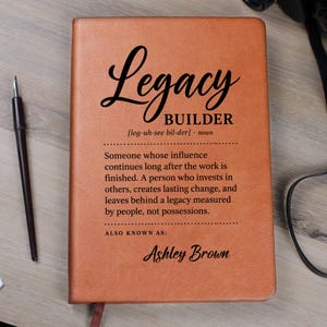 May include: A tan leather-bound journal featuring the black printed text Legacy BUILDER, a phonetic pronunciation, a definition of the term, and the name Ashley Brown at the bottom. A black fountain pen is positioned to the left of the journal.