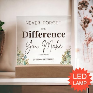 May include: A square clear acrylic plaque set in a rectangular wooden base. The plaque features the printed text "NEVER FORGET THE Difference You Make" with a decorative line and "[CUSTOM TEXT HERE]" below, framed by watercolor-style green foliage.