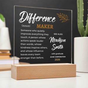May include: A clear acrylic plaque set in a rectangular light wood base. The plaque features white and gold text defining Difference Maker, followed by the name Madison Smith, the phrase with gratitude, ACME CORPORATION, and the year 2026.