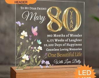Pode incluir: Uma placa de acrílico transparente sobre uma base de madeira retangular com o número 80 em dourado, ilustrações florais com borboletas e texto incluindo To My Dear Friend Mary, 960 Months of Wonder, 4,171 Weeks of Laughter, 29,200 Days of Happiness e With Love, Betty.