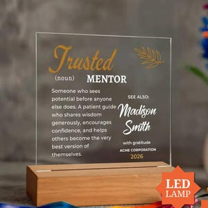 May include: A clear acrylic plaque with gold and white text mounted on a rectangular wooden base. The text reads: Trusted (noun) MENTOR, followed by a definition, the name Madison Smith, ACME CORPORATION, and the year 2026 with a leaf graphic.