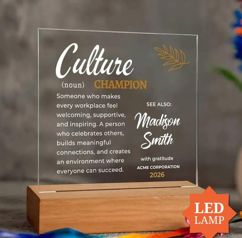 a clear glass plaque with text and a gold leaf design, placed on a wooden stand. The plaque has a definition of "culture" and a name, and it is accompanied by a note about the person named Madison Smith.