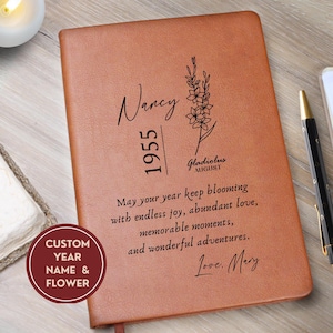 May include: A tan leather-like journal featuring the name Nancy, the year 1955, and a gladiolus flower illustration. The text reads: May your year keep blooming with endless joy, abundant love, memorable moments, and wonderful adventures. Love, Mary.