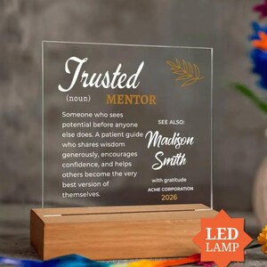 Op de afbeelding: Een doorzichtige acrylplaat in een rechthoekige houten voet. De plaat bevat witte en gouden tekst die een Trusted Mentor definieert, inclusief de naam Madison Smith, ACME CORPORATION en 2026, met een gouden bladgrafiek.