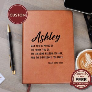 May include: A tan leather-like notebook featuring the name Ashley in black script, followed by the text: MAY YOU BE PROUD OF THE WORK YOU DO, THE AMAZING PERSON YOU ARE, AND THE DIFFERENCE YOU MAKE. TEAM CORP 2025. A black pen sits to the left.