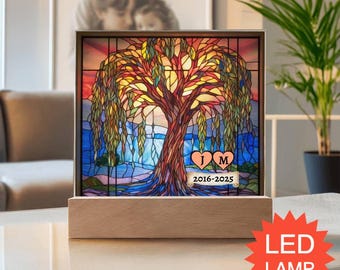 Pode incluir: Uma lâmpada LED quadrada com um design estilo vitral de um salgueiro com folhagem colorida contra um fundo de pôr do sol e água azul. Dois corações cor de pêssego exibem as letras J e M acima de uma faixa com o texto 2016-2025.