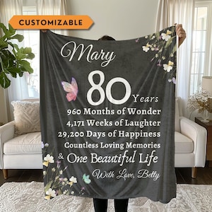 May include: A dark gray throw blanket featuring the name Mary, the number 80, and text detailing 960 months, 4,171 weeks, and 29,200 days. The design includes a colorful butterfly, white and yellow wildflowers, and the closing text With Love, Betty.