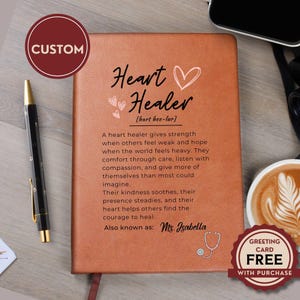 Könnte beinhalten: Ein beigefarbenes Notizbuch in Lederoptik mit dem Text Heart Healer, einem Herzsymbol und einem Text über Stärke und Mitgefühl. Der Einband zeigt den Namen Ms. Isabella und ein Stethoskop-Motiv. Ein schwarzer Stift liegt links neben dem Buch.