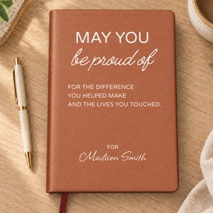 May include: A rectangular brown notebook with white text that reads: MAY YOU be proud of FOR THE DIFFERENCE YOU HELPED MAKE AND THE LIVES YOU TOUCHED. FOR Madison Smith. A white pen with gold accents is positioned to the left of the notebook.