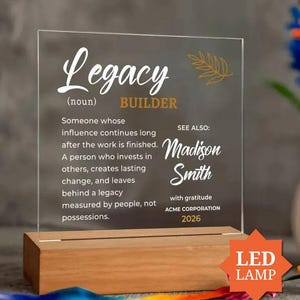 Op de afbeelding: Een doorzichtige acrylplaat met witte en gouden tekst die Legacy (noun) BUILDER leest, inclusief een definitie van de term, de naam Madison Smith, Acme Corporation en het jaar 2026, gemonteerd op een rechthoekige houten voet met LED-lamp.