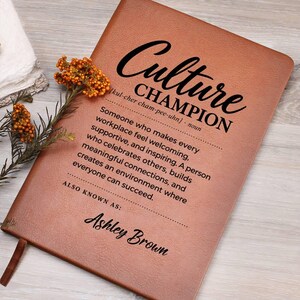 May include: A tan leather-like notebook featuring black printed text that reads Culture Champion, followed by a definition and the name Ashley Brown. A small sprig of dried orange flowers is placed on the left side of the notebook cover.