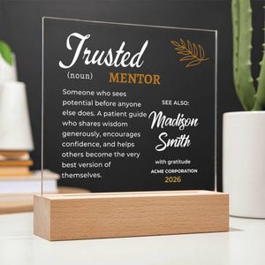 May include: A clear acrylic plaque with a black background featuring white and gold text that defines a trusted mentor. The plaque is set in a rectangular light wood base. The text includes the name Madison Smith and the year 2026.