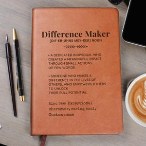 May include: A tan leather-bound notebook featuring the printed text "Difference Maker" followed by a dictionary-style definition and the phrase "Also See: Exceptional character, caring soul, Custom name." Two pens are positioned to the left.