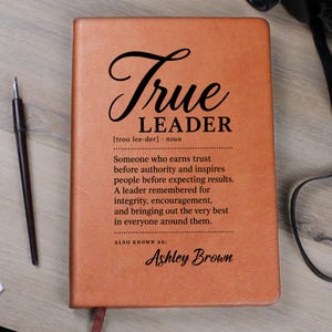 Puede incluir: Un cuaderno de piel sintética color canela con el texto impreso en negro True LEADER, una pronunciación fonética, la definición de un líder y el nombre Ashley Brown en la parte inferior. El cuaderno es rectangular con una cubierta mate.