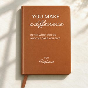 Könnte beinhalten: Ein rechteckiges Notizbuch mit einem glatten, einfarbig braunen Einband. Auf der Vorderseite steht in weißer Schrift: YOU MAKE a difference IN THE WORK YOU DO AND THE CARE YOU GIVE. FOR Stephanie. Ein passendes braunes Lesebändchen ragt unten heraus.