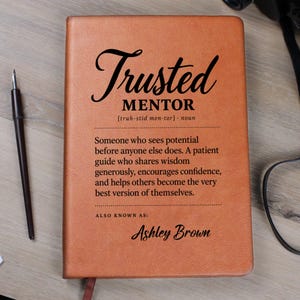 Puede incluir: Un diario con cubierta de cuero color canela que presenta texto impreso en negro que dice Trusted MENTOR, seguido de una definición de mentor y el nombre Ashley Brown. El diario está colocado junto a una pluma estilográfica negra.